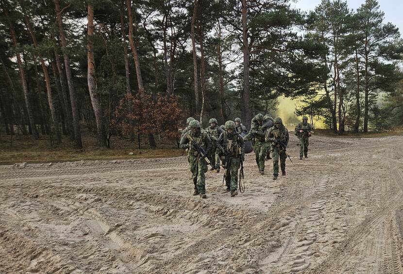Een groepje reservisten loopt tijdens een oefening door een grote zandvlakte met een gewonde op de schouders. Op de achtergrond een grote, gele rookwolk.