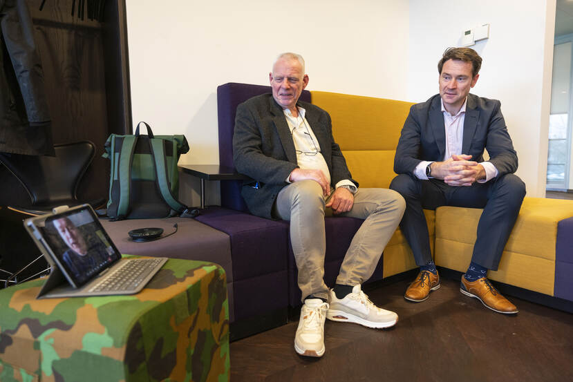 Evert Holtman en Bas Stooker zittend op een bank. Daarnaast een poefje met daarop een iPad, waarop Patrick Benninks gezicht te zien is.