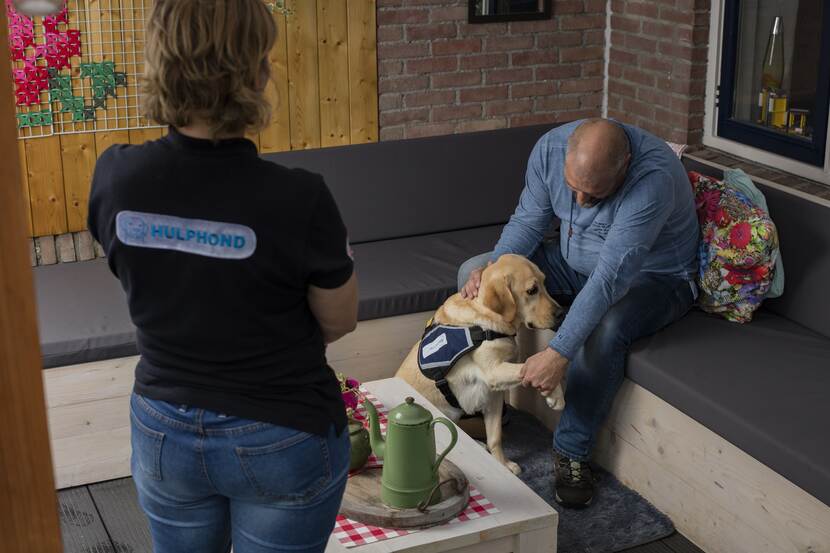 Een man knuffelt op de bank zijn hulphond terwijl een vrouw toekijkt.