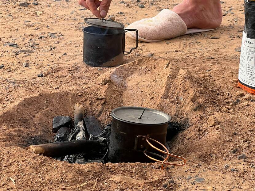 Een gat in het zand met daarin een smeulend vuurtje en op het hout een zwart pannetje.