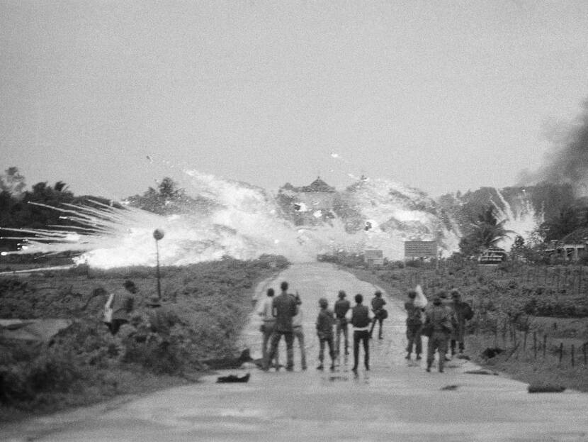 Groepje mensen staat te kijken naar hoe in de verte een bom met veel witte rook inslaat.