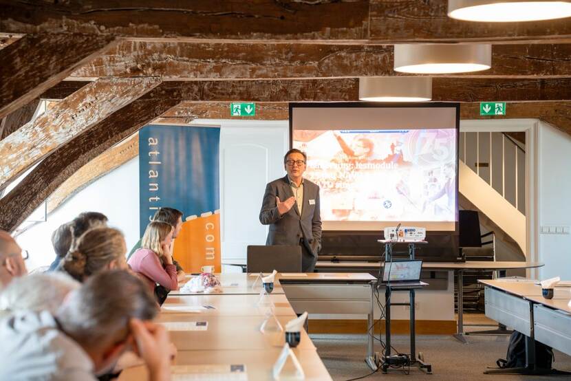 Man staat voor presentatiescherm en spreekt groep deelnemers achter tafels toe