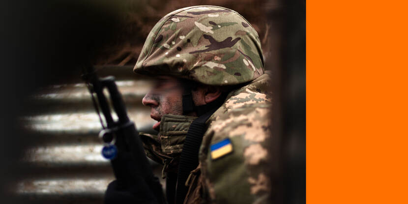 Een Oekraïense militair, met een wapen verticaal in de hand. Hij draagt een helm. Zijn ogen zijn onherkenbaar gemaakt. Er prijkt een blauw-gele Oekraiense vlag op zijn arm.