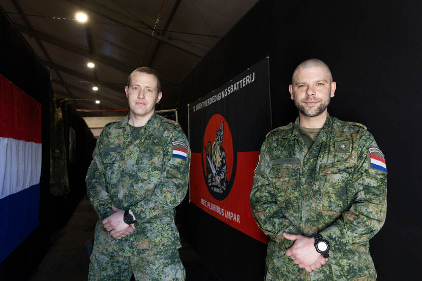 Majoor Jeroen en opperwachtmeester Mikeon poseren naast elkaar, hun handen in elkaar gevouwen voor zich. Achter de twee hangt een vlag van 12 Luchtverdedigingsbatterij.