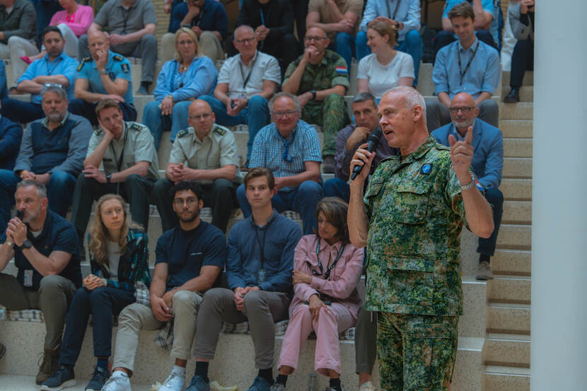 Militair in uniform spreekt met microfoon in de hand volle zaal toe.