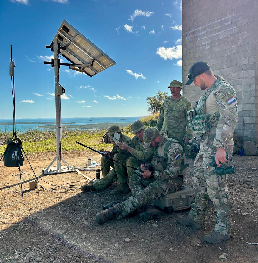 3 militairen zitten op de grond en coördineren via radio en andere apparatuur de vuursteun tijdens een oefening. Achter hen staan 2 militairen die meekijken.