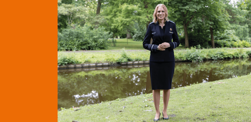 Een vrouwelijke generaal poseert in deftig tenue voor een vijver in bosrijke omgeving.