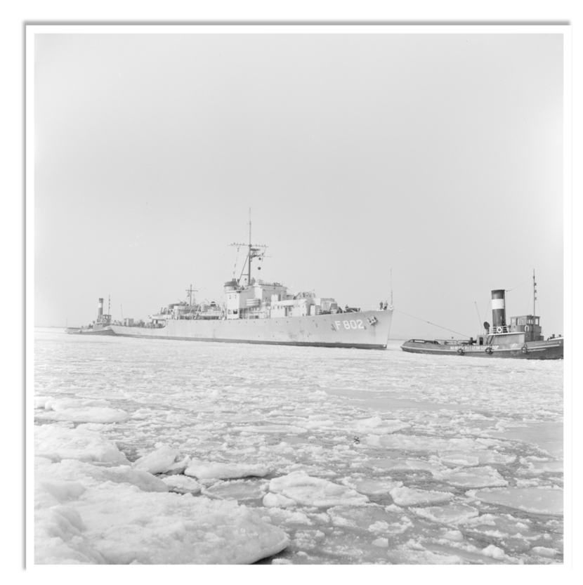 Een groot militair schip met nummer F 802 vaart door ijsvlaktes op zee, begeleid door een kleine sleepboot met een hoge schoorsteen.
