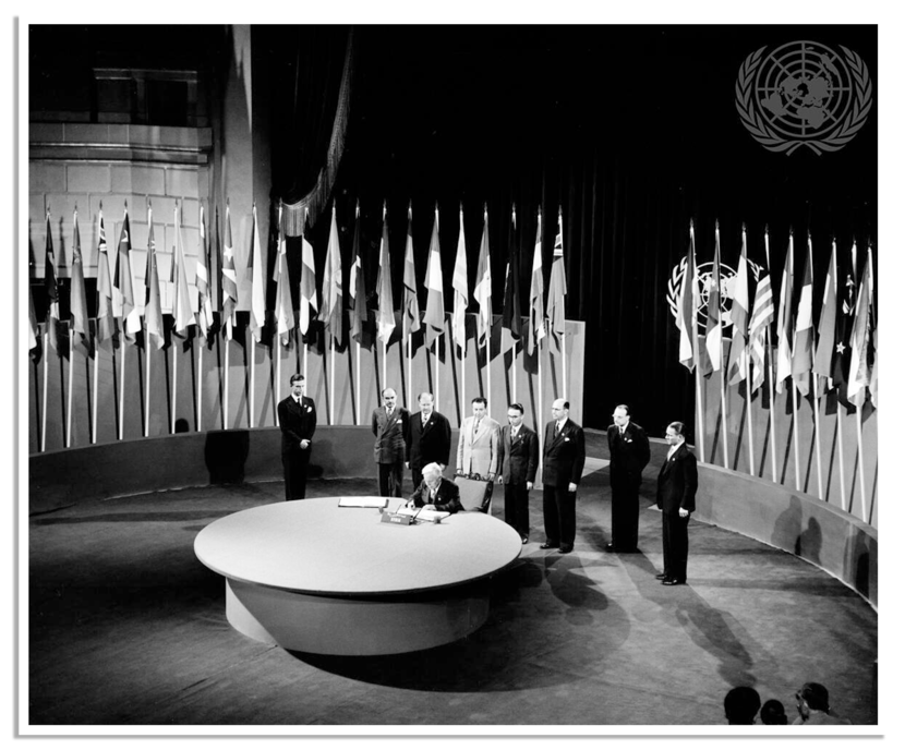Een groep mannen in pak staat plechtig rond een ronde tafel in een grote zaal, omringd door vele vlaggen van verschillende landen, onder het symbool van de Verenigde Naties.