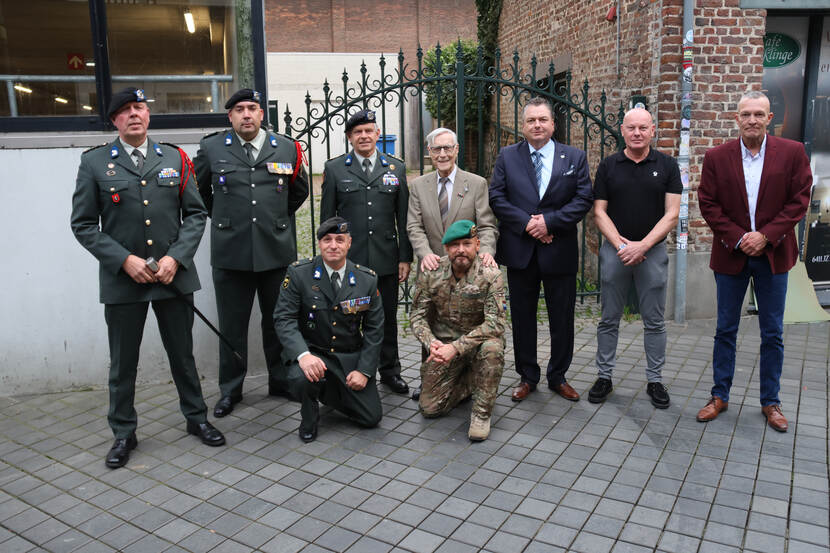 Acht mannen poseren buiten: zes in uniform met medailles, twee in pak. Een man knielt voorin. Achtergrond met bakstenen muur en hek.
