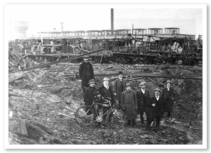 Zeven mannen in pakken en hoeden, staand tussen puin en vernielde spoorwegwagons, met een oude fiets naast hen. Het lijkt op een verwoeste, verlaten industriële omgeving.
