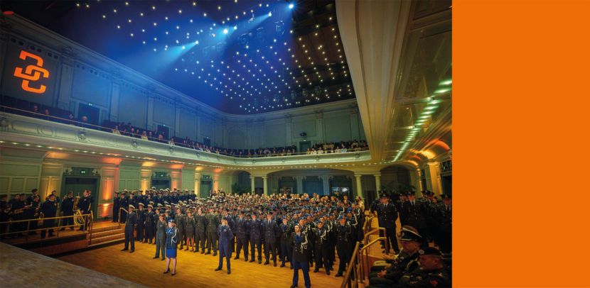 Grote ceremonie in een klassiek theater met honderden militairen in uniform, publiek op balkons en sfeervolle verlichting aan het plafond.