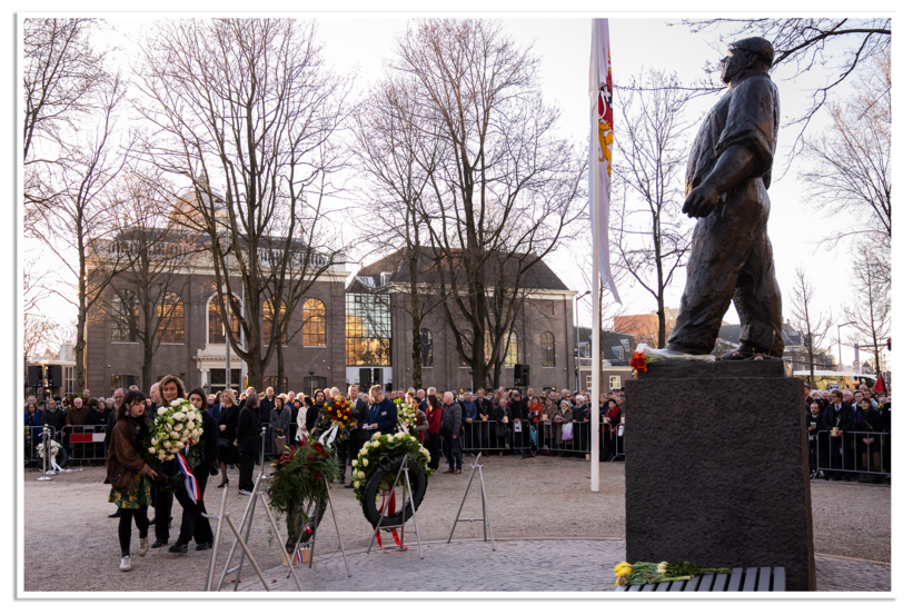 Een herdenkingsplechtigheid bij een standbeeld. Mensen leggen bloemstukken en kransen neer, terwijl een menigte toekijkt bij een gebouw op de achtergrond.