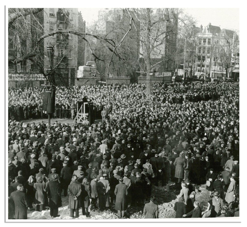Een enorme menigte mensen staat dicht opeengepakt in een openbare ruimte, luisterend naar een toespraak bij een monument, met gebouwen en bomen op de achtergrond.