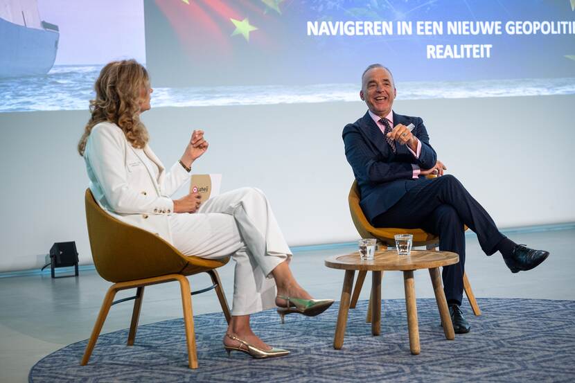 Man en vrouw zitten op podium naast elkaar op een stoel en zijn in gesprek.