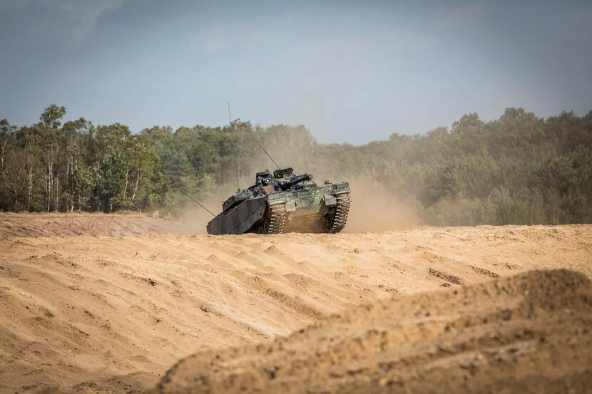 Een rupsvoertuig CV90 verschijnt rijdend boven een zandheuvel.