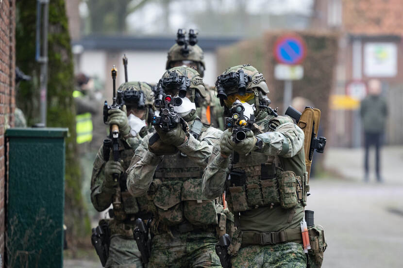 Een groep infanteristen loopt in een treintje langs een gevel. Op de achtergrond kijken burgers toe.