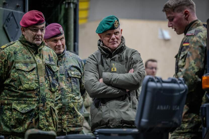 Drie militairen luister naar collega die aan het woord is. Op de voorgrond staat een geopende plastic koffer op tafel.