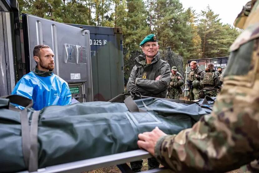 Militair met groene baret kijkt naar collega die brancard verplaatst. Naast hem staat een man met een blauw beschermingspak aan.