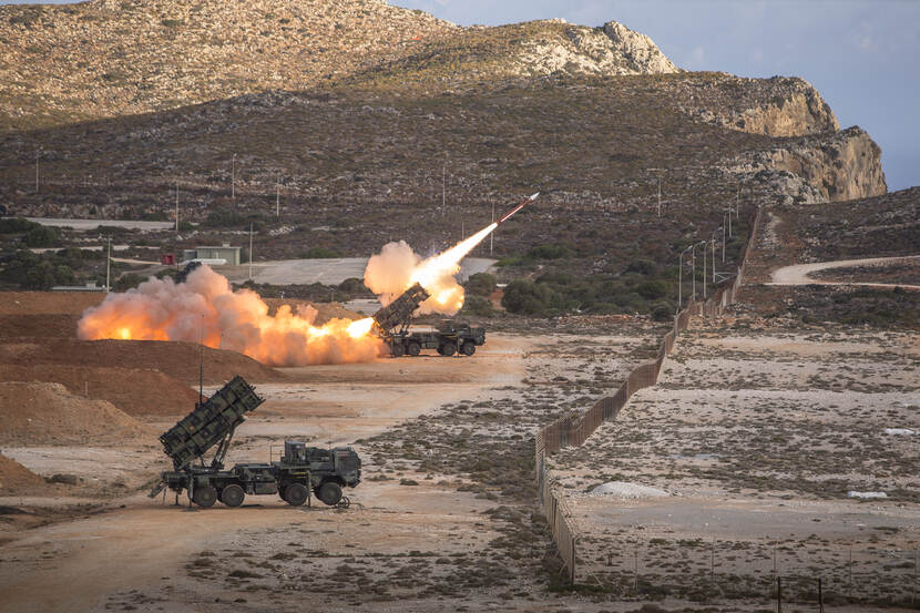 Twee Patriots-lanceerinrichtingen op een zanderig buitenterrein, één ervan vuurt een raket af.