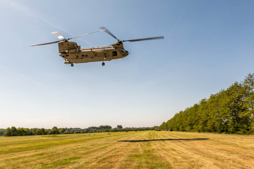 Laag overvliegende Chinook over gras met een de rand een bomenrij.