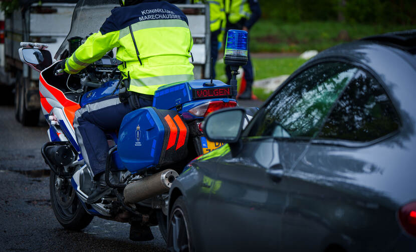 Een motorrijder van de Marechaussee slaat met zwaailichten aan linksaf. Een andere motor staat geparkeerd op het wegdek.