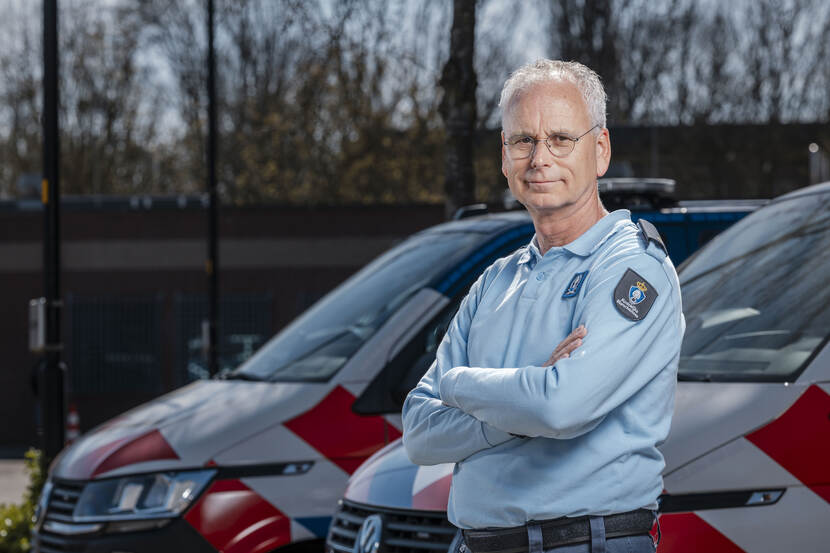 Man met bril in uniform van Marechaussee kijkt vriendelijk en staat met de handen over elkaar voor 2 politiebussen.
