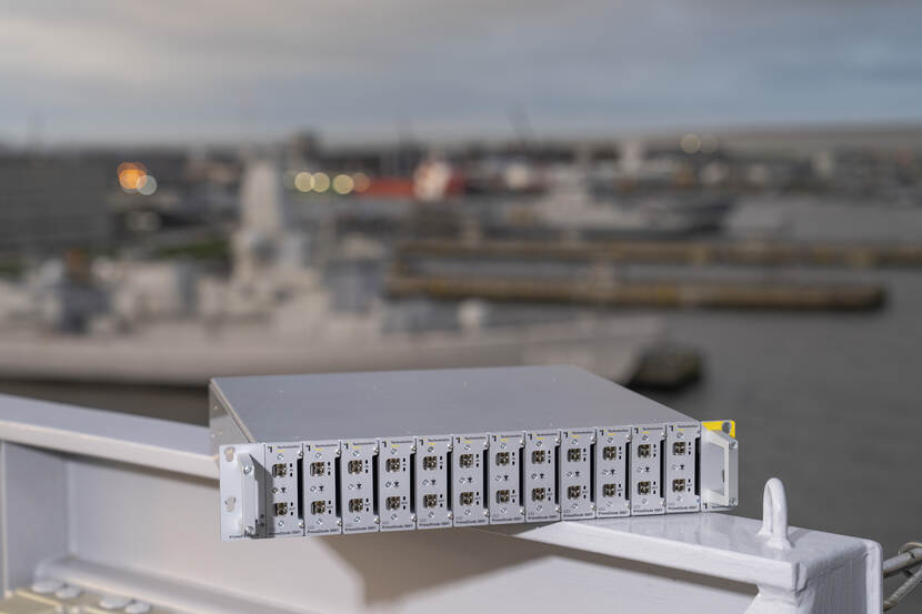 Een los 19 inch serverrek met 12 datadiodes op een hek van het schip met vaag op de achtergrond de marinehaven