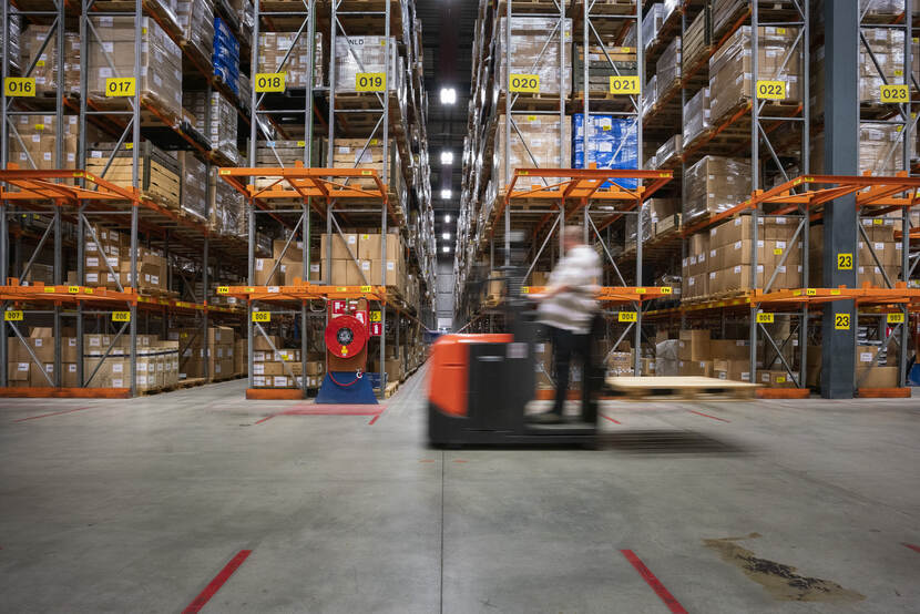 Een medewerker van het KPU passeert de schappen in het magazijn op een steekkarretje en is door de snelheid die hij maakt vaag te zien.