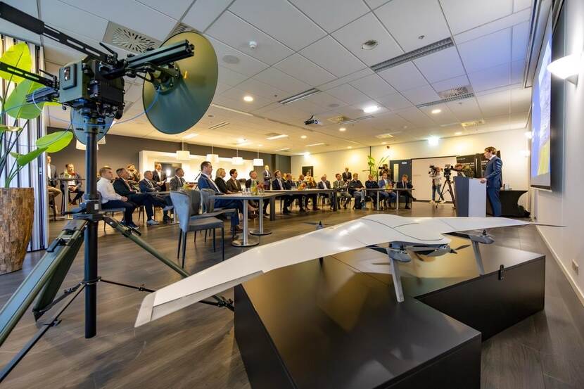 Een gevulde zaal met mensen. De aanwezigen luisteren naar een persoon die door een microfoon spreekt. Op de voorgrond ligt een drone.