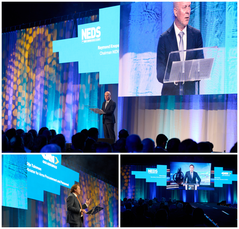 Links en rechts mannen in pak op het podium met blauw licht. Rechts een man met een bril die spreekt vanaf een videoscherm.