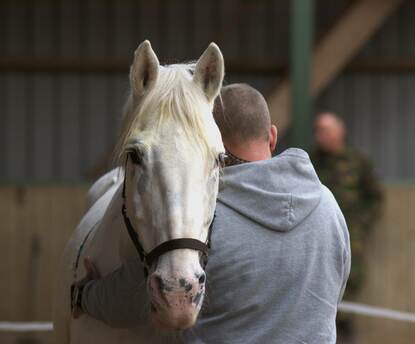 Een paard legt z’n hoofd op de schouder van een grote, gespierde man die met z’n rug naar de camera staat.