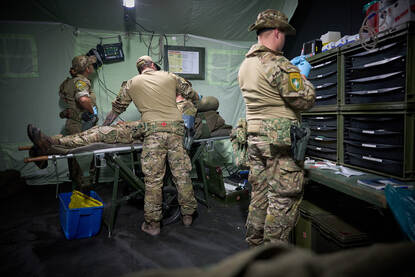 Drie militairen helpen een collega die gewonde speelt in een veldhospitaal. Een van hen heeft een spuit in zijn handen, terwijl de andere twee zich bij de brancard om het slachtoffer bekommeren.