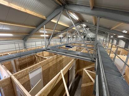 Vanaf een stalen loopburg kijk je op een indeling van gangen en kamers, een soort plattegrond, om te oefenen. De wanden zijn van hout.