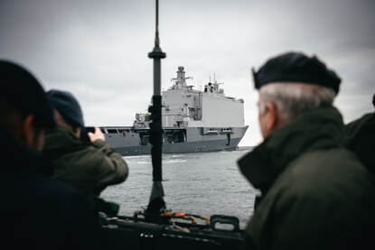 In de verte is amfibisch transportschip Zr.Ms. Johan de Witt op zee te zien. Op de voorgrond staan 2 militairen, van de achterkant bezien. Een van hen maakt een foto van het schip.