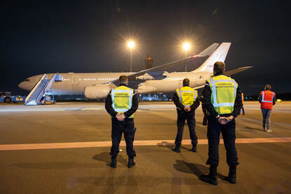 3 personen met een geel hesje van de Marechaussee kijken met de handen op de rug richting een geparkeerd militair vliegtuig.