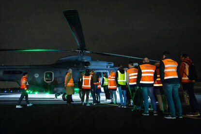 Helikopter landt in het donker op vliegveld. Recht: Groep mensen in oranje hesjes staat in het donker voor helikopter.