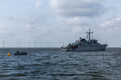 Vanuit een rubber bootje zetten duikers een boei uit, rechts is BNS Lobelia te zien en op de achtergrond de kust van Eemshaven met een aantal windturbines.
