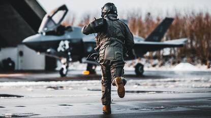 Links stijgt een F-35 in volle vaart op. Rechts rent een vlieger richting zijn gereedstaande kist.