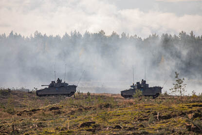Links: Voertuigen en militairen van de battlegroup staan op een veld, met in het midden de vlaggen van de deelnemers. Rechts twee militaire pantservoertuigen rijden door rook en stof op een heuvelachtig terrein, omringd door bos.