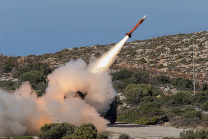 Links: Een raket wordt afgeschoten vanaf militair voertuig met veel rook en vuur, tegen een heuvelachtige, droge en rotsige achtergrond. Rechts: een soldaat die een raket afvuurt vanaf een heuvel met prikkeldraad, terwijl de raket met witte rooksporen door de lucht schiet.
