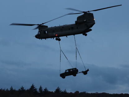 Links een helikopter vliegt in het schemer door de lucht met een voertuig aan een touw er onder. Rechts: Militairen stappen in een helikopter op de heidegrond.