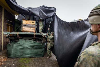 Op beide foto’s zijn militairen druk in de weer met grote zeildoeken om de Scania vrachtwagens te camoufleren.