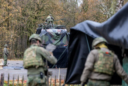 Militairen druk in de weer met zwart zeildoek bij vrachtwagens. Rechts twee militairen op net dak van een vrachtwagencabine.