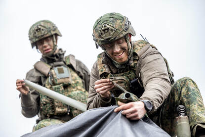 Militairen druk in de weer met zwart zeildoek bij vrachtwagens. Rechts twee militairen op net dak van een vrachtwagencabine.