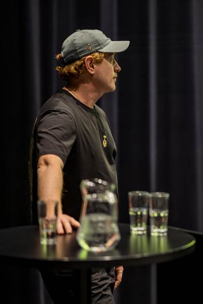 Denys met een hand leunend op een tafeltje voor zich uit starend.