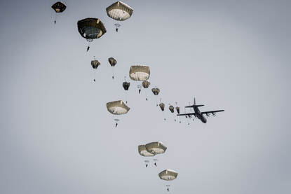 Zo’n 20 parachutisten springen uit een vliegtuig en hangen naast elkaar in de lucht.