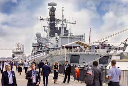 Een fregat aan de kade van London Excel, met een helikopter op het dek. Veel mensen kijken en lopen rond.
