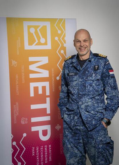 Militair staat voor banner en kijkt lachend in de camera.