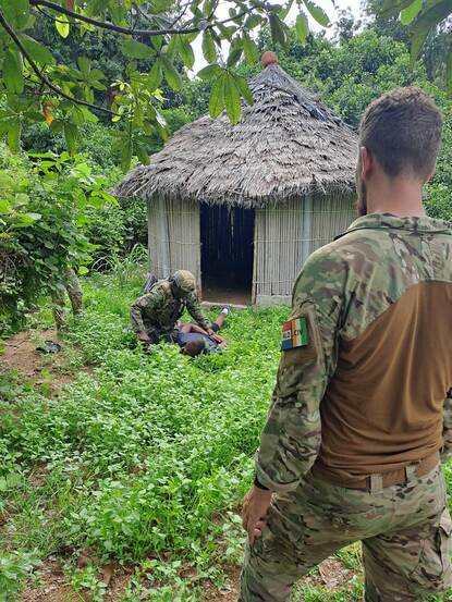 Een Nederlandse militair van de rug bezien, met voor hem een Ivoriaanse militair geknield boven een oefenslachtoffer voor een huisje.